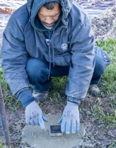 City worker smoothing freshly poured concrete around a purple martin pole base with gloved hands