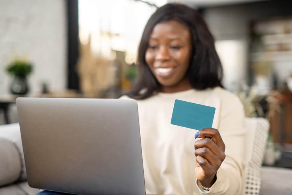 Cheerful young multiracial woman shopping online with credit card and laptop while sitting on sofa at home,